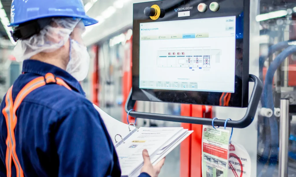 A man wearing a hard hat and safety glasses is focused on a computer screen in a work environment.