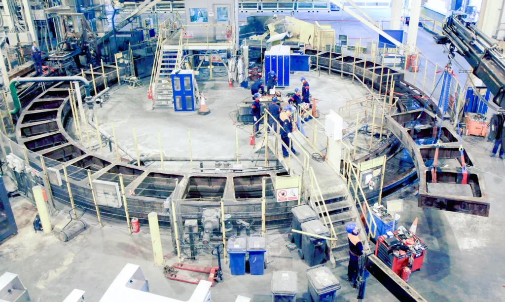 A circular metal structure is under construction inside a factory, with workers and machinery visible in the background.