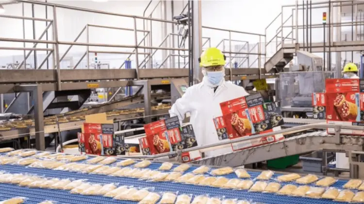 A worker in a white coat stands by a conveyor belt loaded with food items, overseeing the production process.