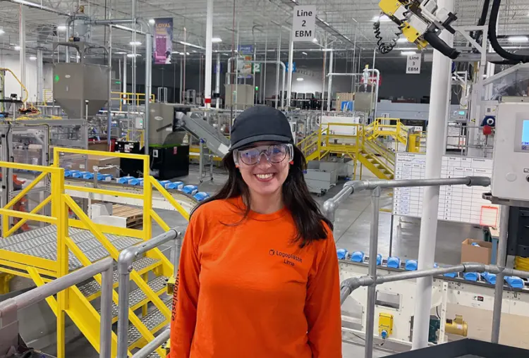 A woman in an orange shirt and glasses stands in a factory, surrounded by machinery and equipment.