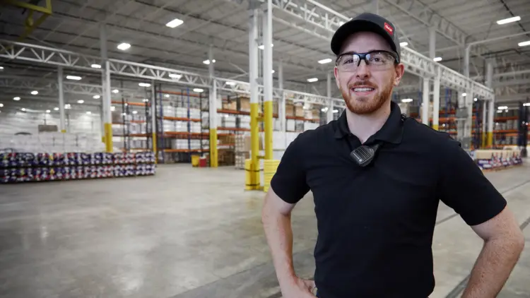 A man wearing a black shirt and glasses stands in a warehouse, surrounded by shelves and boxes.