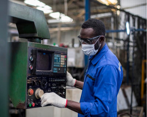 A man in a blue shirt and face mask operates a machine in a workshop setting.