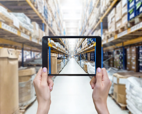 ipad-techcrunch-500×400 A person in a warehouse holds up a tablet, showcasing information or data to an unseen audience.