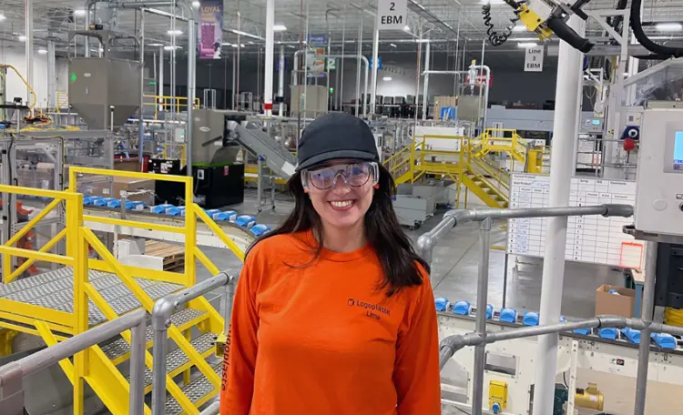 A woman in an orange shirt and glasses stands in a factory, surrounded by machinery and equipment.